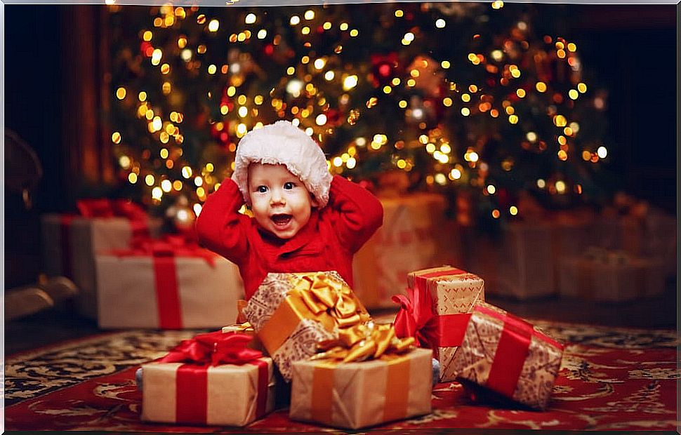 Baby opening his Christmas presents under the tree decorated with lights.